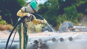 Male worker performing industrial sand blasting outdoors, wearing full protective gear and using a high-pressure hose to clean metal pipes, with dust and abrasive media visibly in the air. 