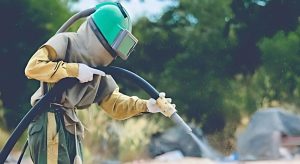Worker in full protective gear using a sandblasting machine outdoors, demonstrating proper technique and safety measures for sandblasting plastic components with controlled pressure and specialized media. 