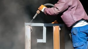 Worker using a sandblasting nozzle to clean or strip paint from a wooden or metal frame, surrounded by dust and wearing protective gear, demonstrating a process similar to sand blasting paint off wood for surface restoration. 