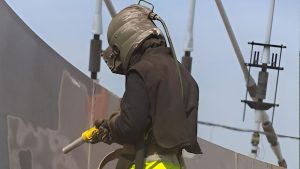 Technician engaged in industrial sand blasting, wearing full protective gear while cleaning a large metal surface to remove old coatings and prepare for refinishing.