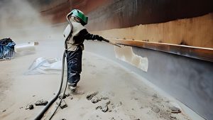 Worker in protective gear performing surface cleaning on a large metal structure, demonstrating the sandblasting definition in action with abrasive material visibly removing coatings.