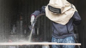 Worker using protective gear while performing abrasive blasting on a surface, demonstrating professional service quality for those searching “sandblasting near me.”