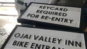 Two painted wood signs with dimensional black text show examples of carving letters in wood, featuring phrases like "KEYCARD REQUIRED FOR RE-ENTRY" and "OJAI VALLEY INN BIKE ENTRANCE," showcasing clean routed lettering on a textured background.