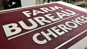 Close-up of a dimensional HDU sign for Farm Bureau Cherokee featuring raised white lettering on a textured maroon background. This example showcases carving letters in wood-style signage with clean edges, bold typography, and precise routing for professional sign applications. 