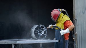 A worker in protective gear sandblasting a wheel, removing dirt and debris to prepare it for refinishing. Sandblasting wheels in progress.