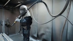 A worker wearing protective sandblasting gear, operating a sandblasting machine in a controlled environment, demonstrating local sandblasting services.