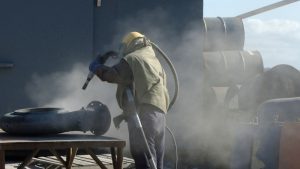 Worker using a sandblasting machine to clean and prepare aluminum rims, with dust clouds rising in the process of restoration and customization.