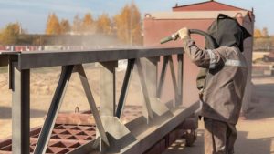Worker performing local sandblasting on a metal structure outdoors, using a hose to apply abrasive material for surface preparation.