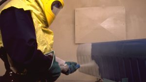 Worker in protective gear using abrasive blasting equipment to strip paint from a metal surface, showing small parts sandblasting services near me.