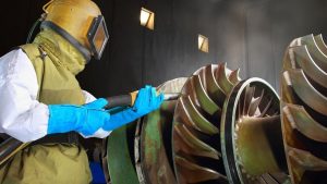 Worker sandblasting aluminum rims with protective gear in an industrial setting.