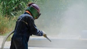 Worker in protective gear using industrial equipment for sandblasting wood, creating a smooth and clean surface for restoration.