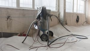 A sandblasting machine set up in a room with sand covering the floor, showing equipment used for surface preparation.