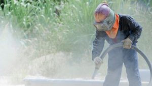 Worker in full protective gear using a blasting hose for sandblasting wood, clearing dust and debris from the surface outdoors.
