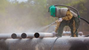 Worker wearing protective gear while sandblasting metal pipes to remove rust and surface impurities.
