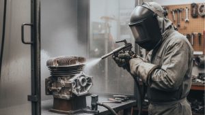 Technician wearing protective gear using a sandblaster to clean a metal engine part, illustrating how does a sandblaster work in removing rust and surface contaminants.