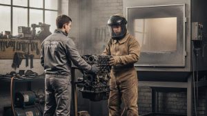 Two workers in a workshop, one in protective gear holding a car engine block, preparing it for sandblasting.