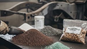 Different types of blasting media, including walnut shells, glass beads, and garnet, displayed on a table. These materials are commonly used in sandblasting for car frame restoration, showcasing the best media for sandblasting car frame.