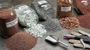 Sandblasting media displayed in labeled jars, including aluminum oxide grit, glass beads, steel grit, and walnut shells, showing different abrasive types used for surface preparation and cleaning.