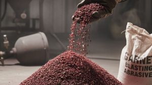 Hand pouring garnet sandblasting media from a bag, with a pile of the abrasive material on the ground in an industrial setting.