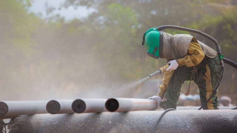 Worker sandblasting metal pipes with protective gear, removing rust and surface contaminants during industrial surface preparation