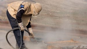 A worker sandblasting steel with protective gear and a hose.