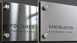 Comparison of polished stainless steel and sandblasted stainless steel finishes on mounted metal signage panels with visible texture differences.