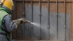 Worker using sandblasting media for steel to remove rust and corrosion from a metal gate before surface restoration and repainting.