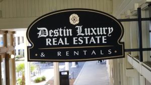 HDU sign for Destin Luxury Real Estate & Rentals with white lettering on a black background, mounted on a metal bracket above a walkway.